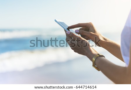 Hipster girl hold on smart phone gadget in sand coastline, mock up of blank screen. Traveler using in female hand mobile on background seascape horizon. Tourist look on blue sun ocean, lifestyle 
