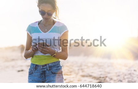 Hipster girl photograph on smart phone gadget in sand coastline beach, mock up of blank screen. Traveler hold and using in female hand mobile on background seascape horizon. Tourist look on sun ocean