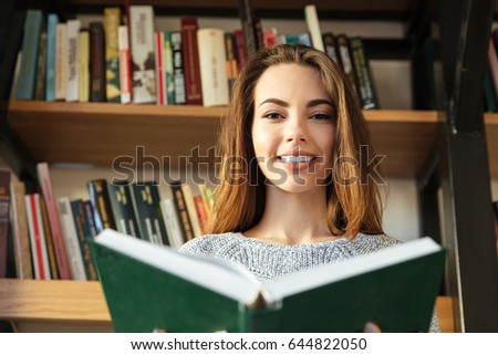 Photo of young happy woman student sitting in library reading book. Looking at camera.