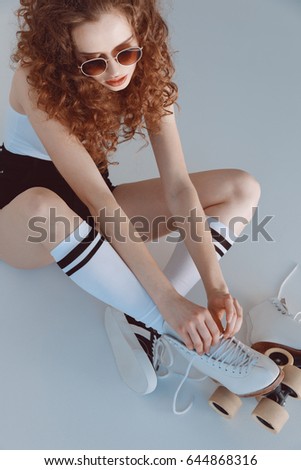 High angle view of hipster girl in sunglasses wearing roller skates sitting isolated on grey 