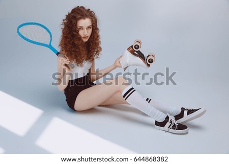 Attractive stylish hipster girl sitting and holding tennis racket and roller skate 