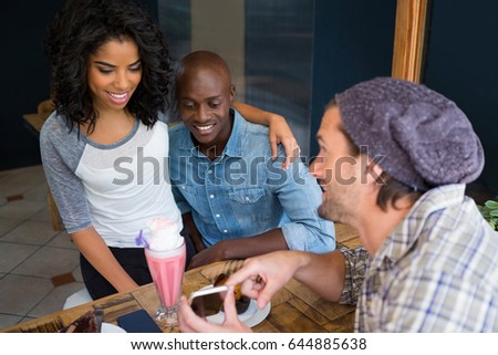 Young man showing mobile phone to couple at table in coffee shop