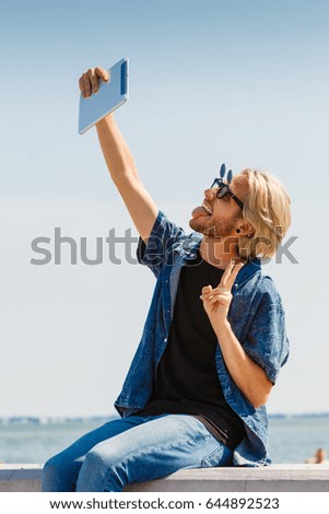 Contacts, technology, self-esteem concept. Young blonde man on vacations using his tablet to take cool selfie shot showing peace symbol