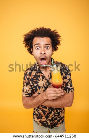 Picture of young shocked african man standing isolated over yellow background. Looking at camera drinking cocktail.