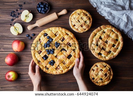 Homemade organic apple pies hold female hands on dark wooden kitchen table with raising, bluberry, rolling pin and apples. Traditional dessert on Independence Day. Flat lay bakery food background. Royalty-Free Stock Photo #644924074