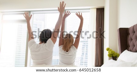 Back view of young happy couple in white t-shirts waking up in the morning, sitting on bed, stretching in cozy bedroom, looking through window. Horizontal photo banner for website header design 