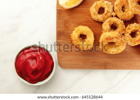 An overhead photo of calamari rings with a slice of lemon, and tomato sauce, on a white marble texture with a place for text