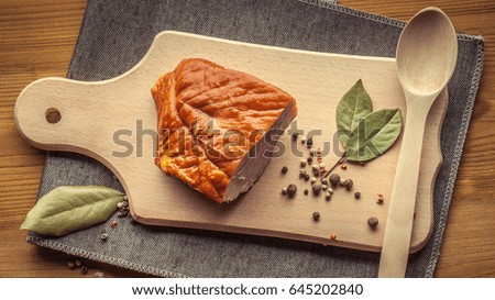 A piece of smoked meat, a mixture of dry spices of peppers, a bay leaf on a kitchen cutting board, a wooden spoon on a brown wooden background