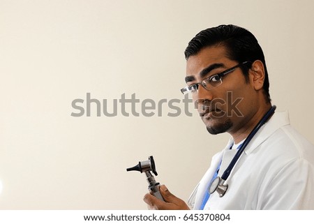 Young handsome doctor wearing blue scrubs and a white lab coat holding otoscope looking to the side 