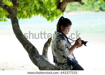 young asia woman sitting on timber under tree and using camera for take photo. have sea are background. this image for people,photographer and nature concept