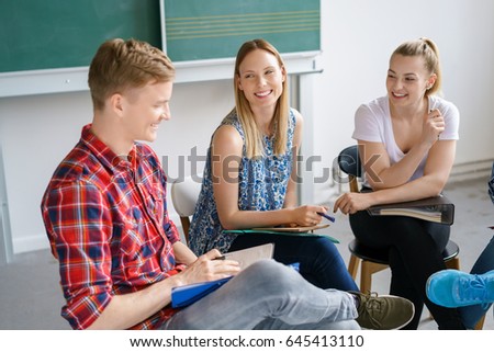 Smiling diverse young students having a group discussion in the classroom in front of the chalkboard sitting in a circle on chairs
