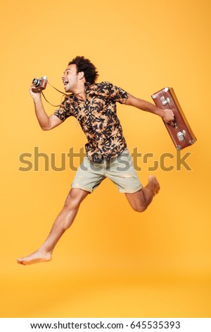 Full length portrait of a happy african man in summer clothes jumping and taking photo with retro camera isolated over yellow background