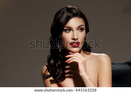 Young pretty beautiful woman in red long evening dress with makeup, red lips and classical hollywood waves hairstyle sitting in director's chair at film set with two cinema lights.