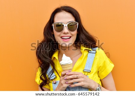 Beautiful  funny young hipster teen girl eating ice cream cone. laughs happy. Bright casual wear, denim shorts. Orange  background, urban style, sunglasses