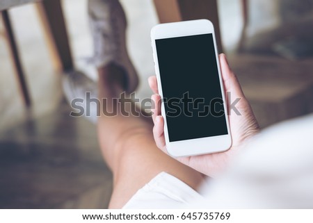 Mockup image of woman's hand holding white mobile phone with blank screen on thigh with wooden floor background in modern