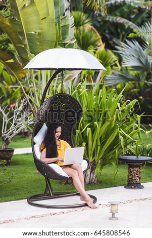Young successful woman works as freelancer on-line in outdoor garden. Brunette in yellow dress with silver laptop. Internet work in travel