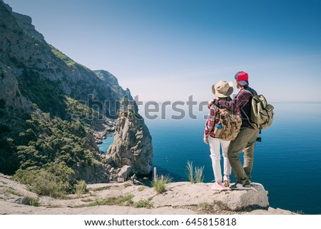 Couple Travelers Man and Woman in the Mountains look at the Sea. Young family hikers with backpacks outdoors. Freedom and active lifestyle concept.