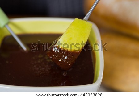 Close up of a delicious apple in a metal stick covered with chocolate fondue inside of a white bowl on wooden table