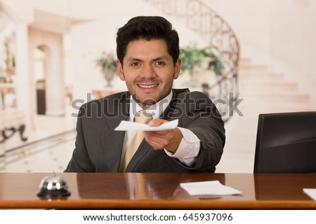 Happy smiling receptionist in hotel giving key to guest and papers to sign, hotel background
