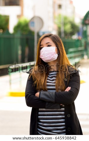 Young woman with protective mask on the street in the city with air pollution with her arms crossed, city background