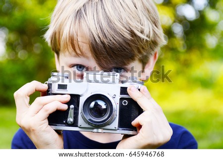 Little child blond boy with an old camera shooting outdoor. Kid taking a photo using a vintage retro film camera. Green summer grass lawn background.