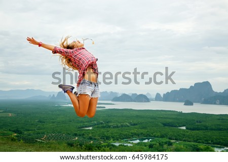 Lifestyle image of happy young traveling woman jumping carefree on nature background. In the mountains. Wearing stylish short shorts and checkered shirt. Freedom and happiness concept. In motion Royalty-Free Stock Photo #645984175