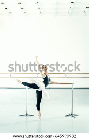Pretty young ballerine doing stretching ballet exercise in bright ballet studio. Toned image.