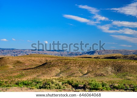Green bushes, desert hills with mountains in the distance under blue sky with white clouds.
