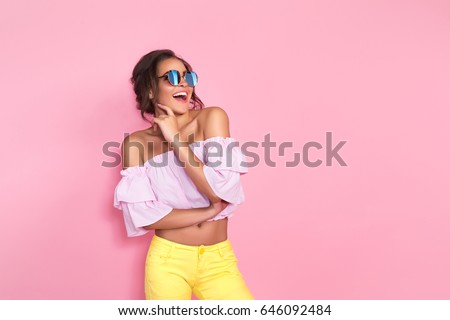 Beautiful girl in yellow jeans and pink shirt wearing sunglasses posing, screaming, smiling on pink background in studio. 