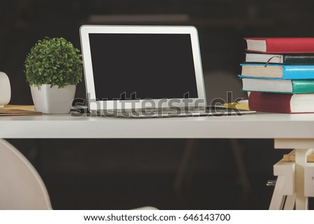 Horizontal shot of creative workplace with blank laptop, stacked books, decorative plant and other items. Mock up