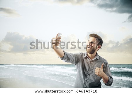 Portrait of cheerful young guy showing thumbs up and taking selfie at the beach