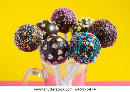 Chocolate cake pops in glass, decorated with colorfull confectionery sprinkles on a yellow background. Close-up. Picture for a menu or a confectionery catalog.