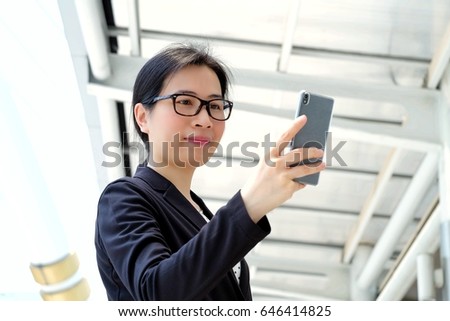 Beautiful young asian business woman standing in front of office building in business area and use smart phone.