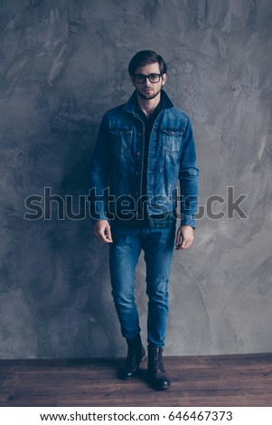 Autumn. Young serious bearded guy in casual jeans outfit is standing on a concrete wall`s background, wearing glasses and look pensive Royalty-Free Stock Photo #646467373