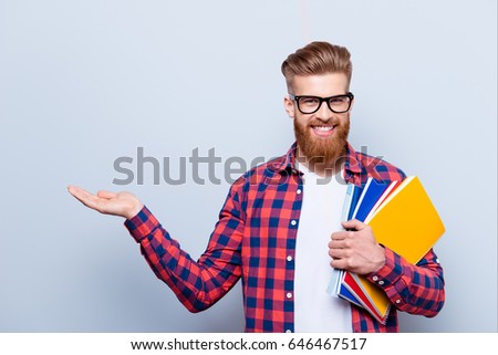 Cheerful young nerdy red bearded student is standing with books on pure background in glasses and casual bright outfit, holding something - copyspace