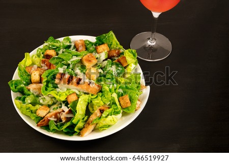 A photo of a plate of chicken Caesar salad, with a glass of rose wine, on a black background with a place for text