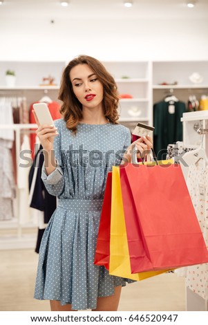 Thoughtful woman holding bags and credit card while using smartphone in shop