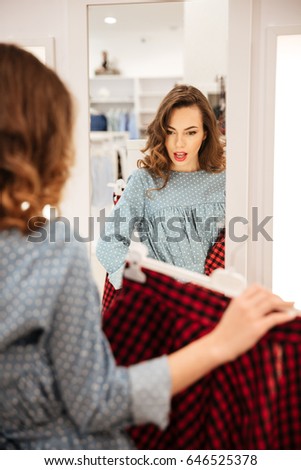 Photo of serious young woman shopper in blue dress looking at mirror in shop choosing clothes. Looking aside.