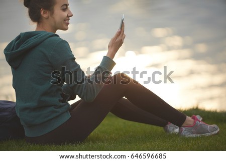 Portrait of beautiful happy girl in sportswear with small urban backpack on the shore of a lake or sea. woman makes selfie photo or video call.