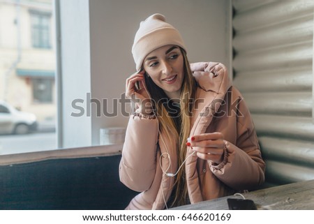 The lovely girl puts on earphones in cafe. On her a pink cap and a jacket. She looks aside and grins. The girl has beautiful long hair.