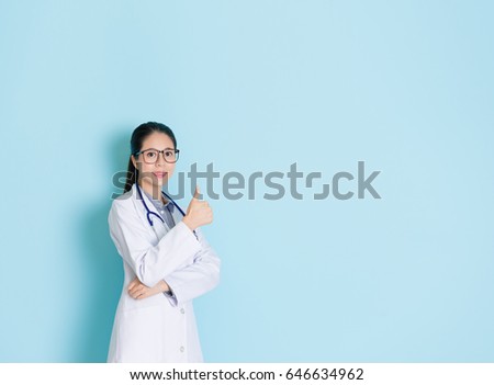professional attractive lab female medical doctor showing finger thumb up gesture and face to camera smiling standing in blue wall background.