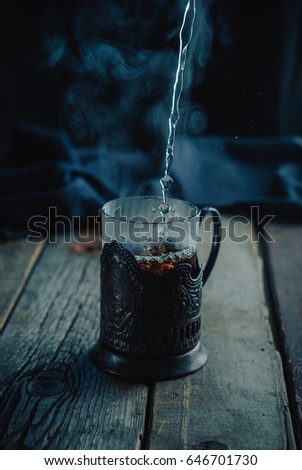 Very hot tea with steam, pouring into a glass in a cup holder on a wooden vintage rustic table. Black background. Dark and moody. Toned picture. Close up, selective focus