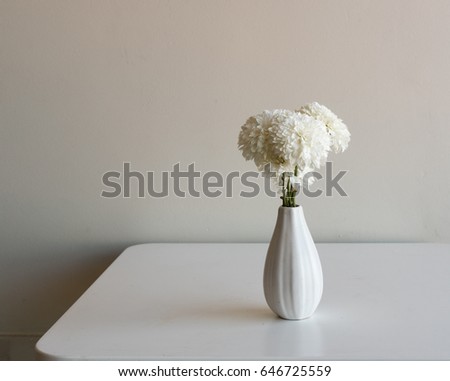 Wilting white chrysanthemums in small white vase on edge of table against neutral background