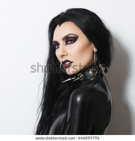 Beautiful dominant brunette vamp fetish goth mistress evil girl in black latex dress and bdsm spiky leather collar posing on white backgroung