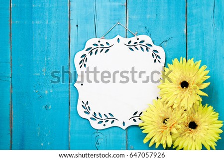 Blank wood sign with floral border and yellow flowers hanging on antique rustic teal blue wooden door; springtime, Mothers Day, family, Memorial Day holiday background with white copy space