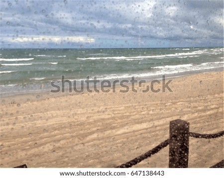Sandy beach, blue sea and cloudy sky through the window glass with rain drops on it.