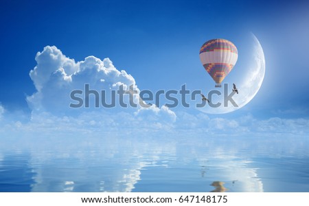 Idyllic heavenly picture - colorful hot air balloon, two seagulls flying in blue sky with white clouds and crescent above serene sea. Dream come true concept.Elements of this image furnished by NASA Royalty-Free Stock Photo #647148175