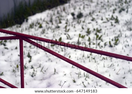 Water droplets on a red, scarlet fence against the background of white May snow and green grass. For wallpaper, advertising, design, covers, background, textures.