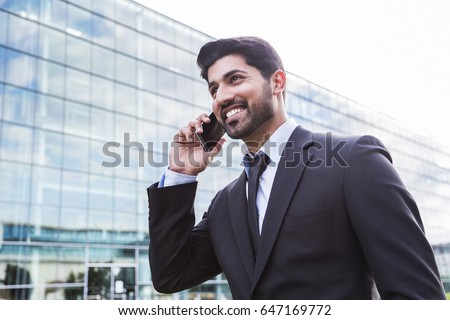 Arabic serious smiling happy successful businessman or worker in black suit with tie and shirt with beard calling with his phone near his ear standing in front of an office building. Royalty-Free Stock Photo #647169772