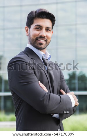 Arabic serious smiling happy successful businessman or worker in black suit with tie and shirt with beard and arms crossed on chest standing in front of an office building. Royalty-Free Stock Photo #647170399